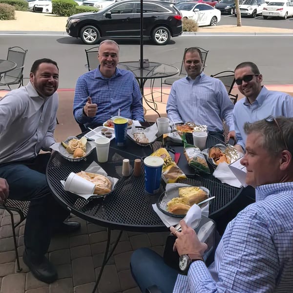 Five men enjoying deli sandwiches and drinks at an outdoor patio table at Rinaldi's Italian Deli.