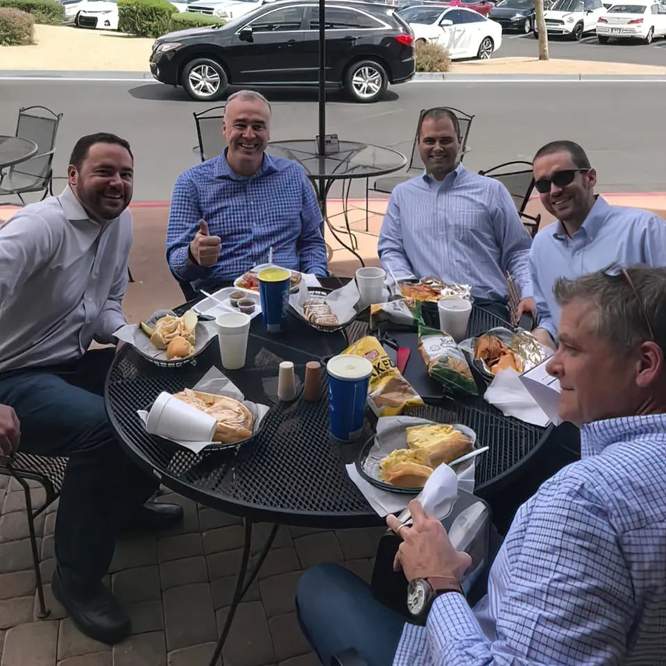 Five men enjoying deli sandwiches and drinks at an outdoor patio table at Rinaldi's Italian Deli.