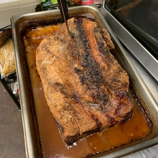 A large, roasted brisket with a dark, crispy crust sits in a metal pan with juices.
