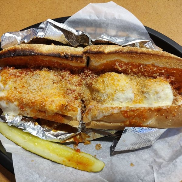 A Rinaldi's Italian Deli meatball sub, cut in half, with melted cheese and sauce, served with a pickle.