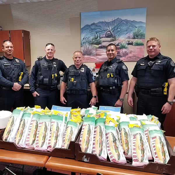 Five police officers standing behind a table laden with Rinaldi's Italian Deli submarine sandwiches and chips.