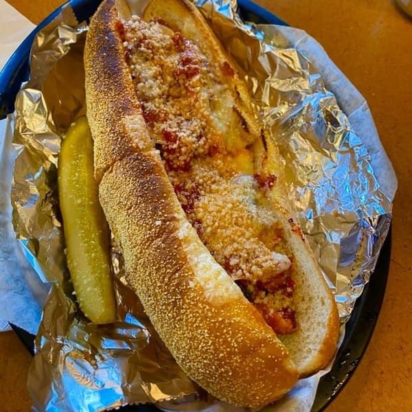 Meatball sub with melted cheese and sauce, served with a pickle spear on foil.