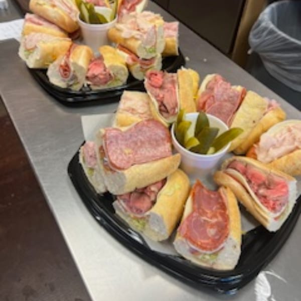 Two platters of Rinaldi's Italian Deli sandwiches, cut in half, with pickles.