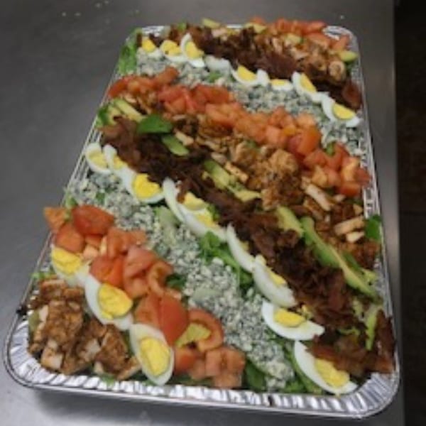 A large catering tray of Cobb salad with chicken, bacon, avocado, tomatoes, and hard-boiled eggs.